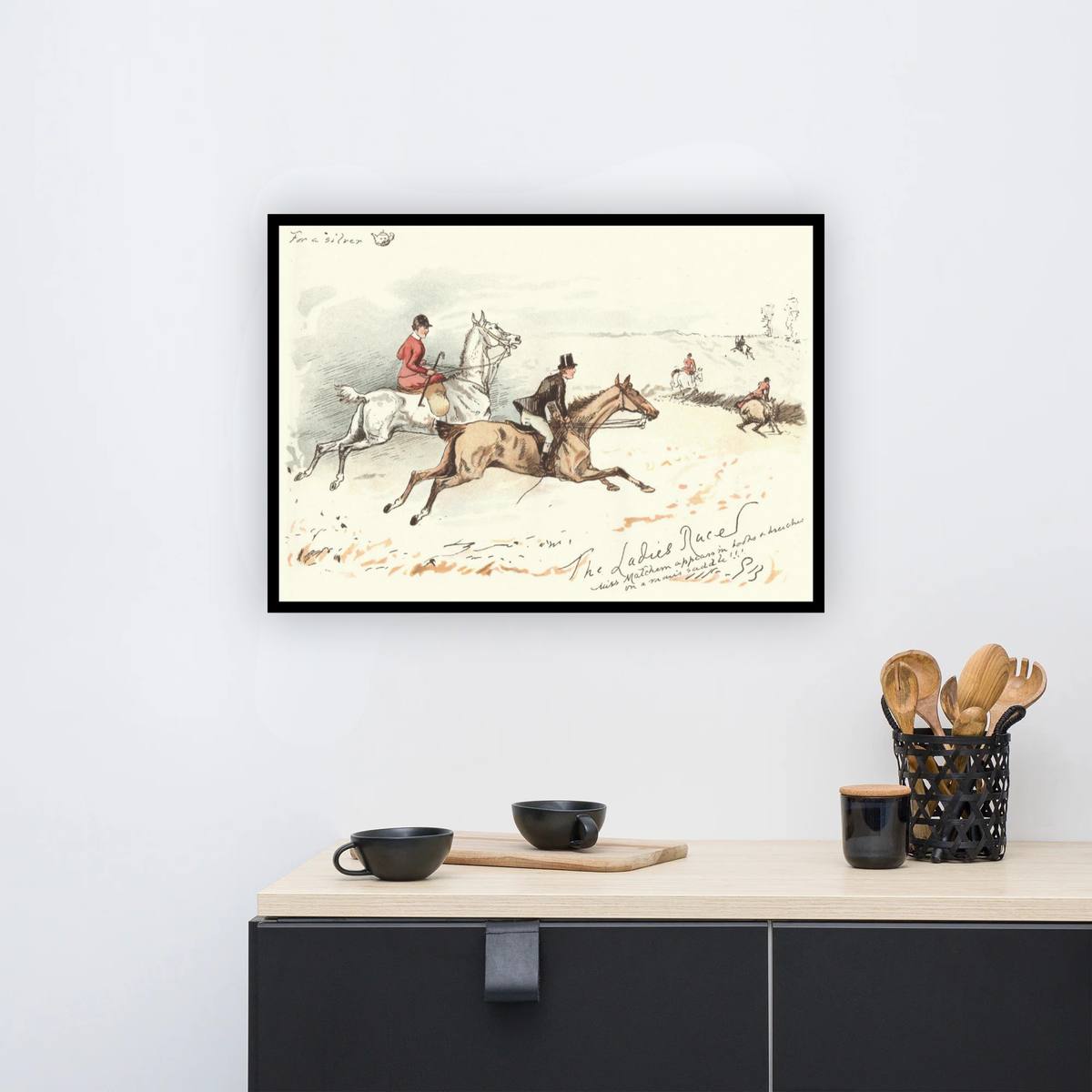 A framed vintage equestrian art print hangs on a white wall above a kitchen counter with black mugs and wooden utensils.