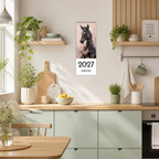 A vertical 2027 wall calendar featuring a black horse portrait, hanging on a light beige kitchen wall above mint green cabinets. The scene is a bright, modern kitchen with wooden countertops, potted plants, and a hanging green mug.