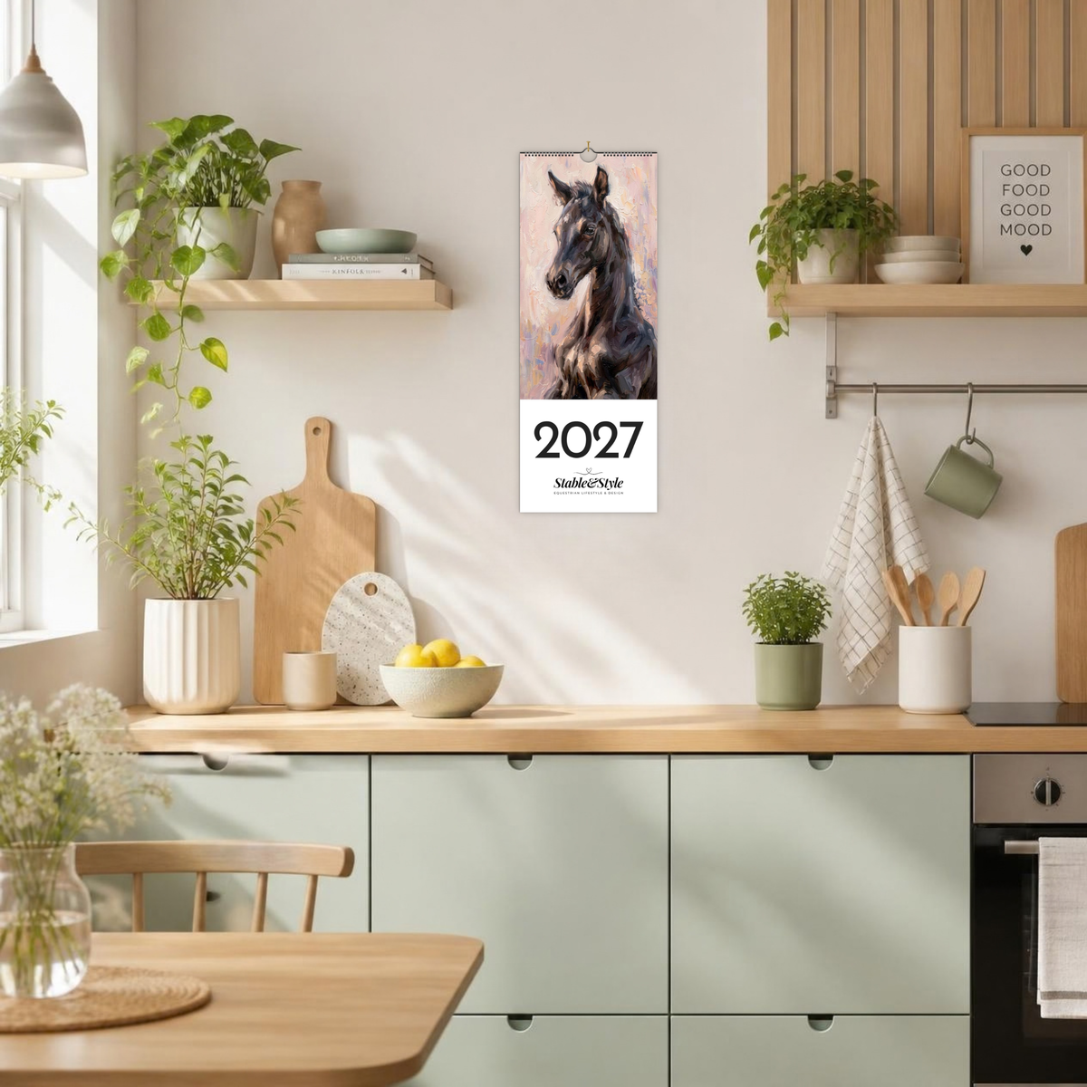 A vertical 2027 wall calendar featuring a black horse portrait, hanging on a light beige kitchen wall above mint green cabinets. The scene is a bright, modern kitchen with wooden countertops, potted plants, and a hanging green mug.