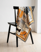 A patterned silk scarf with horse illustrations and geometric borders in orange, beige, black, and gray, draped over a black wooden chair in a minimalist room with light wood floors.