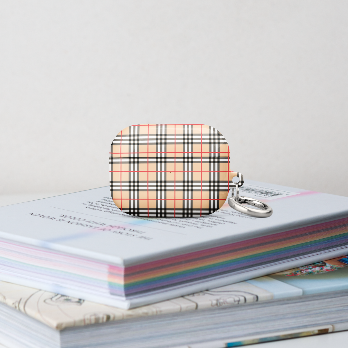 An airpods case featuring a creme, red, black, and white plaid pattern, resting on top of a stack of pastel-coloured books. A silver metal key ring is attached to the side.
