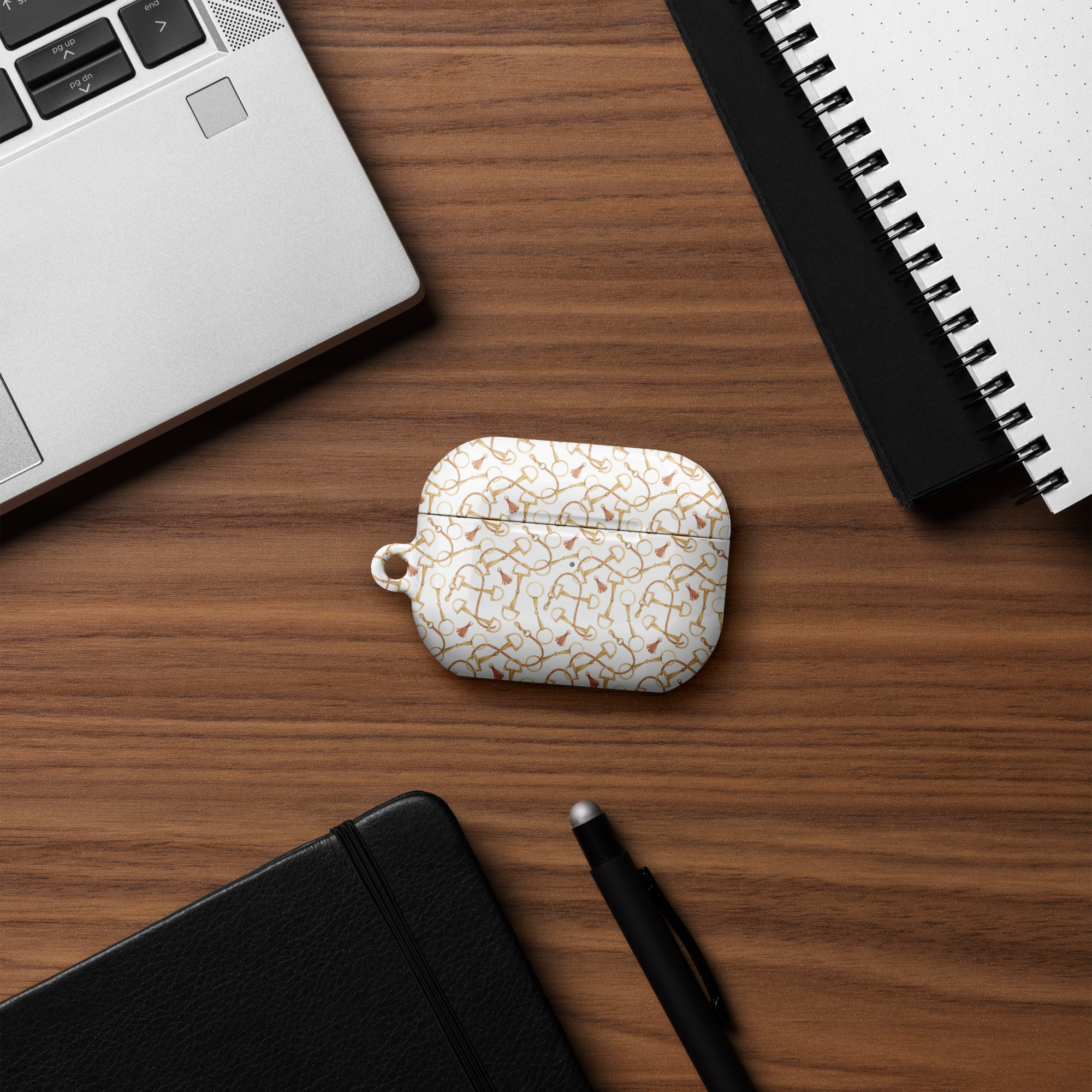 White earbud case with gold pattern on a wooden desk with laptop, notebook, and pen.