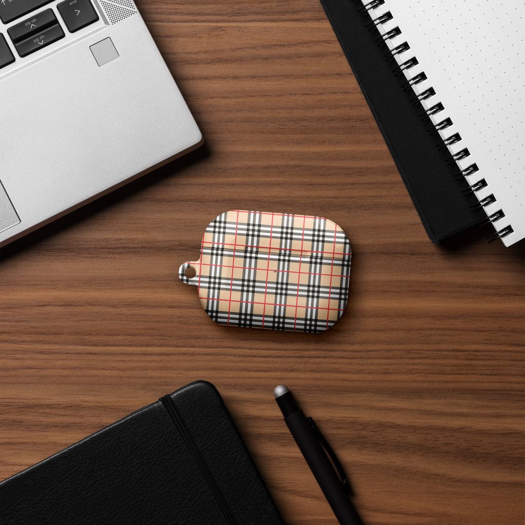 An airpods case featuring a creme, red, black, and white plaid pattern, placed on a wooden desk alongside a laptop corner, spiral notebook, closed black notebook, and a pen.