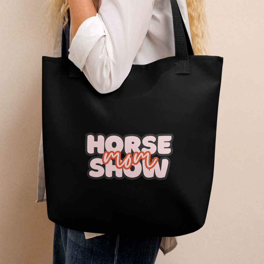 A woman with a black tote bag, with the words "HORSE SHOW MOM" in white, orange  and pink. The scene is photographed against a plain beige wall.th