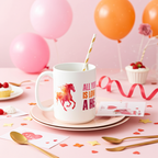 White mug with a horse design and text on a pink tablecloth with balloons and cupcakes.